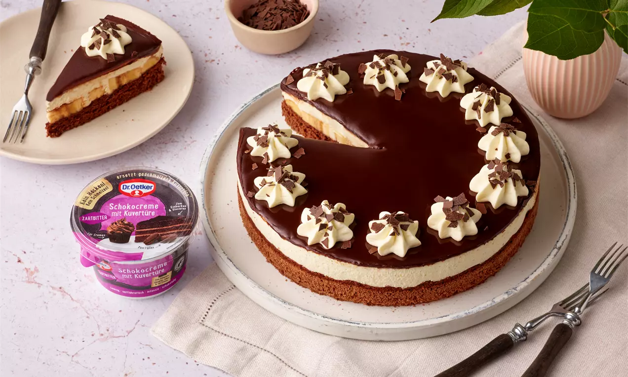 Schoko-Bananen-Torte mit glänzender Schokoglasur, Sahnetupfen und Bananenstücken, serviert auf einem hellen Teller mit Besteck und Deko