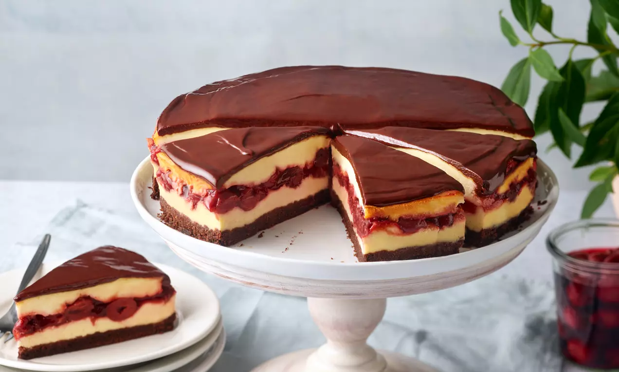 Schwarzwälder Käsetorte mit dunklem Schokoguss, hellgelber Creme und roter Kirschschicht auf weißer Tortenplatte mit angeschnittenem Stück