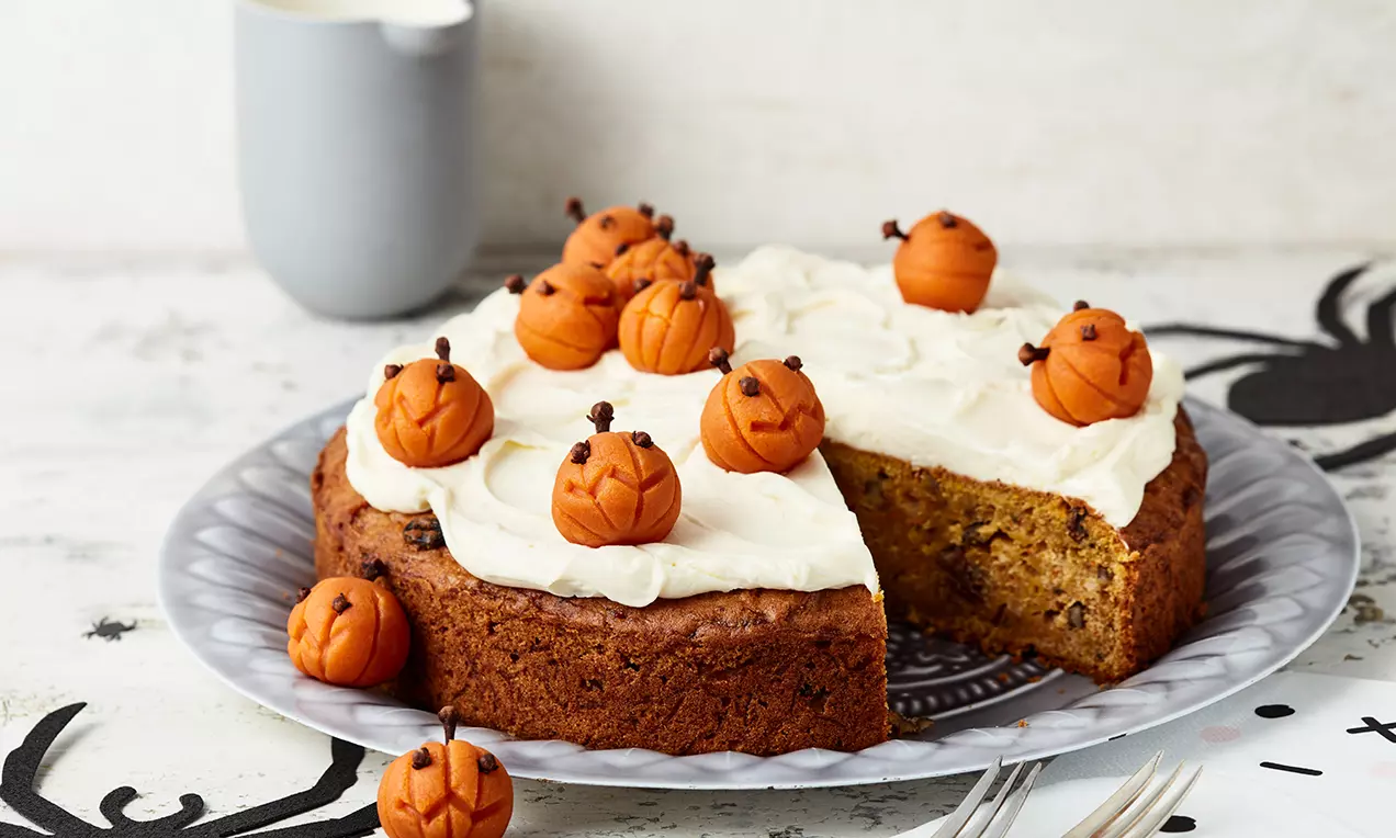 Saftiger Kürbiskuchen mit weißer Creme, gespickt mit kleinen orangefarbenen Marzipankürbissen, auf einer gemusterten grauen Platte angerichtet