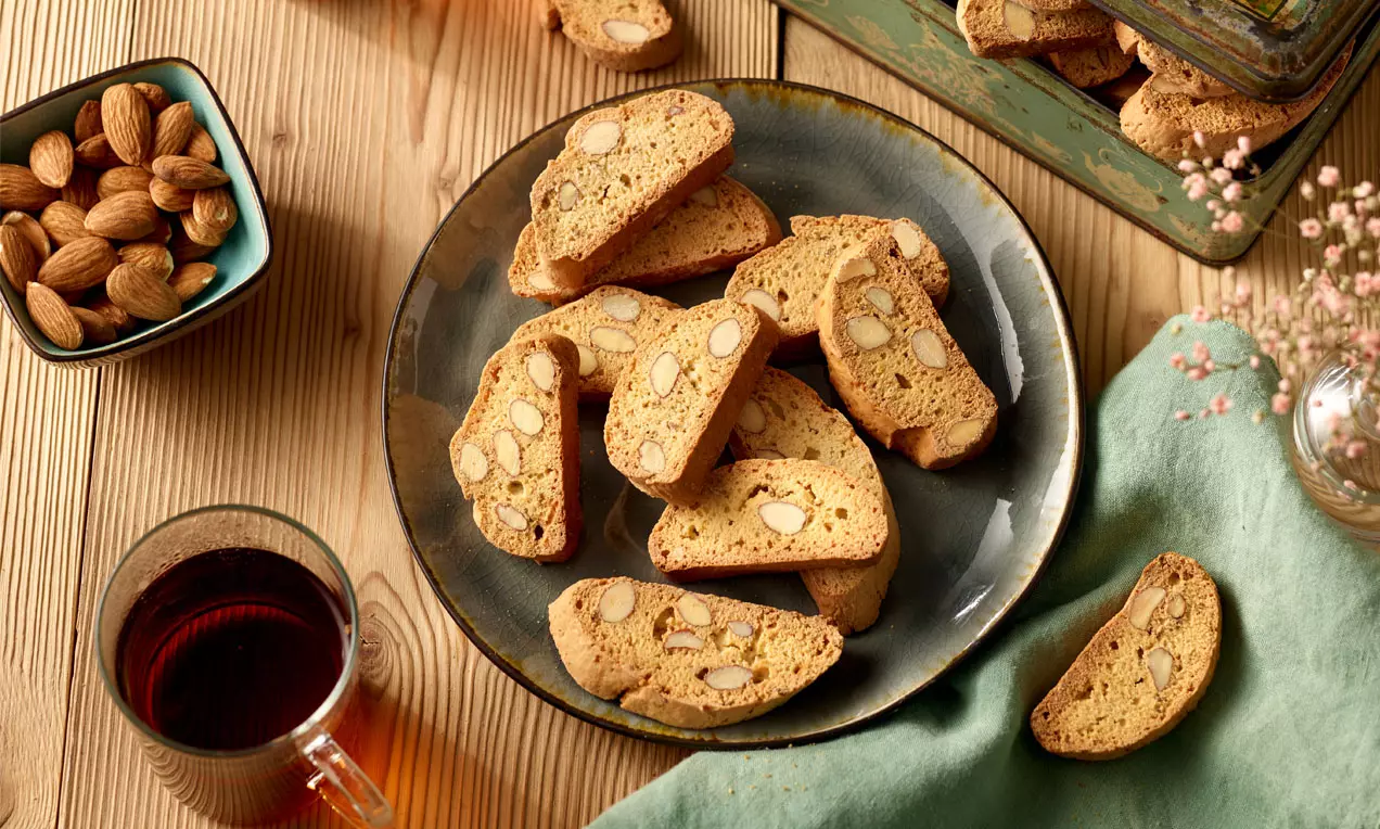 Knusprige, goldbraune Cantuccini mit Mandeln auf einem dunklen Teller mit grünem Tuch, neben Tee, Mandeln und einer Blechdose drapiert