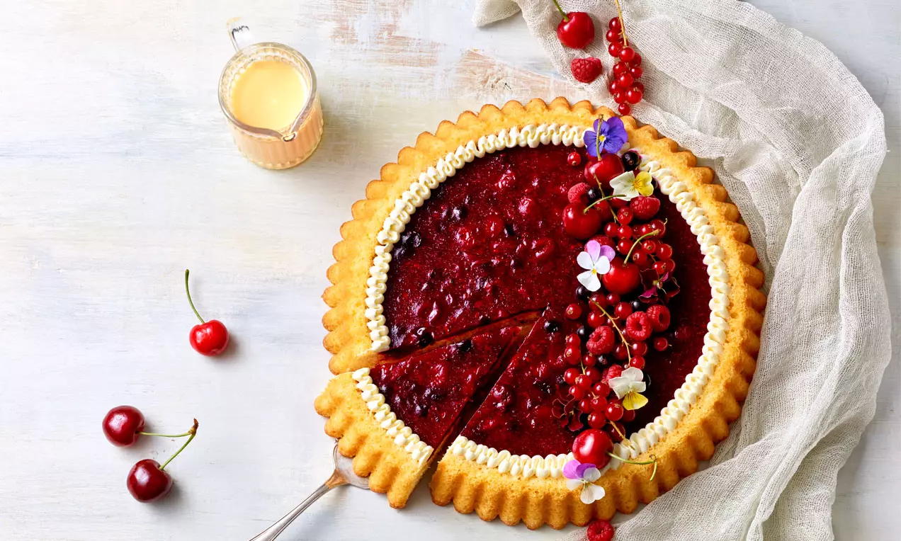 Runder Kuchen mit goldgelbem Rand, glänzend roter Beerenfüllung, Sahnebordüre, Blüten und Beeren auf hellgrauem Tuch angerichtet