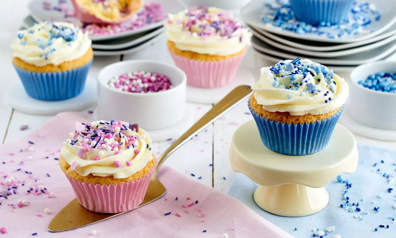 Bunte Cupcakes mit cremiger weißer Haube und farbigem Zuckerdekor, in rosa und blauen Förmchen, auf Tellern und Pastelltüchern angerichtet