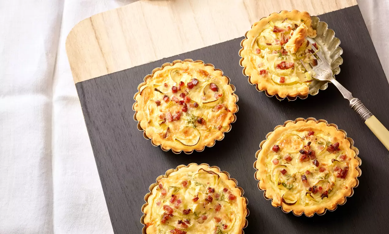Kleine, goldbraune Pizzatörtchen mit welligem Rand, saftiger Füllung aus Käse, Lauch und Schinkenwürfeln auf dunklem Holzbrett angerichtet