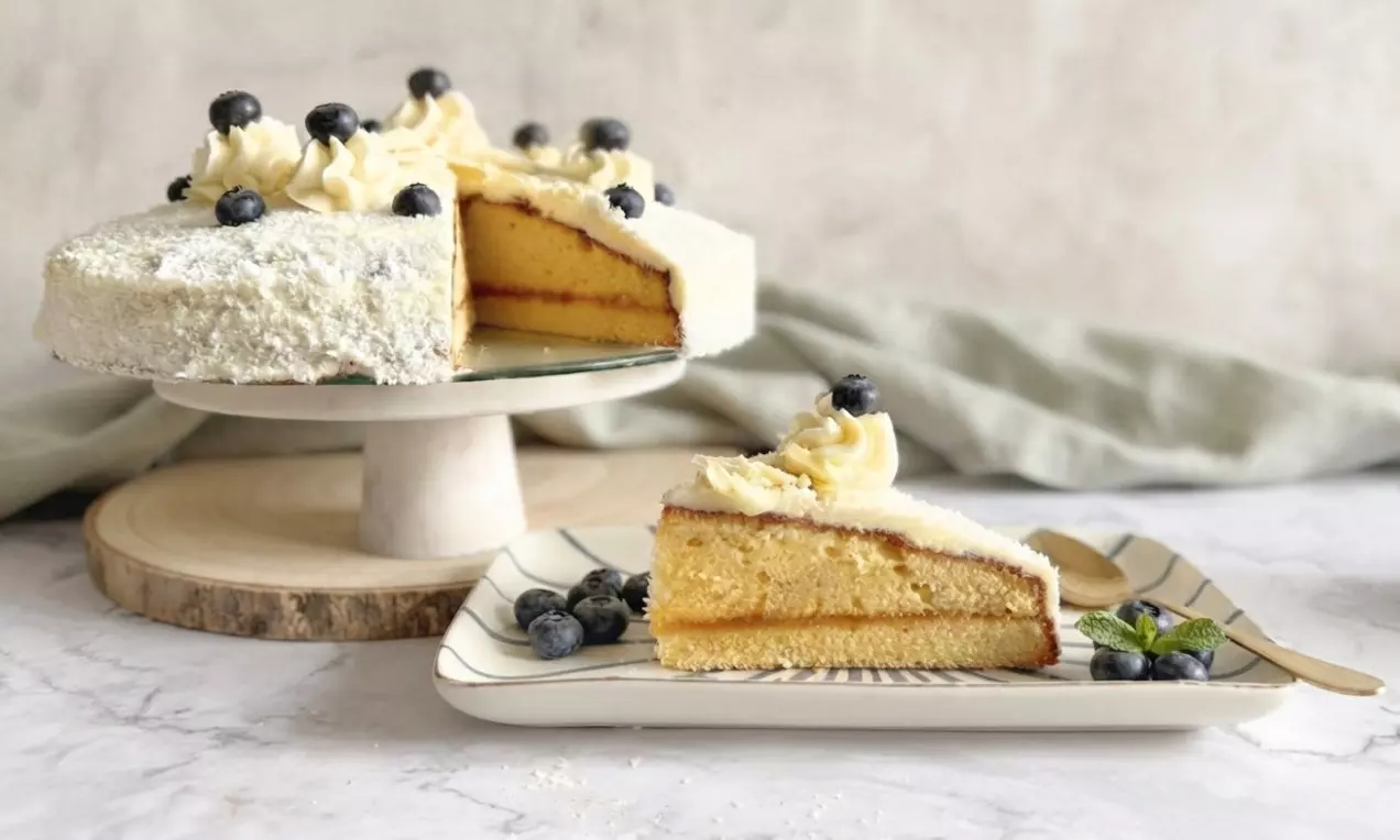 Weiße Sachertorte mit heller Glasur, dekoriert mit Sahnehäubchen und Blaubeeren, ein angeschnittener Kuchen auf einer Platte und ein Stück auf einem Teller