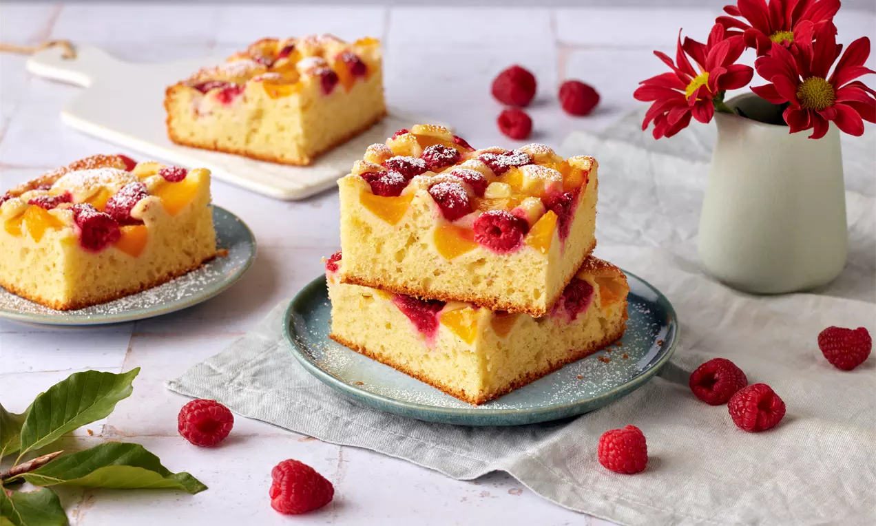 Zwei quadratische Stücke heller Rührkuchen mit saftigen Pfirsichspalten und Himbeeren, bestäubt mit Puderzucker, auf einem Teller angerichtet