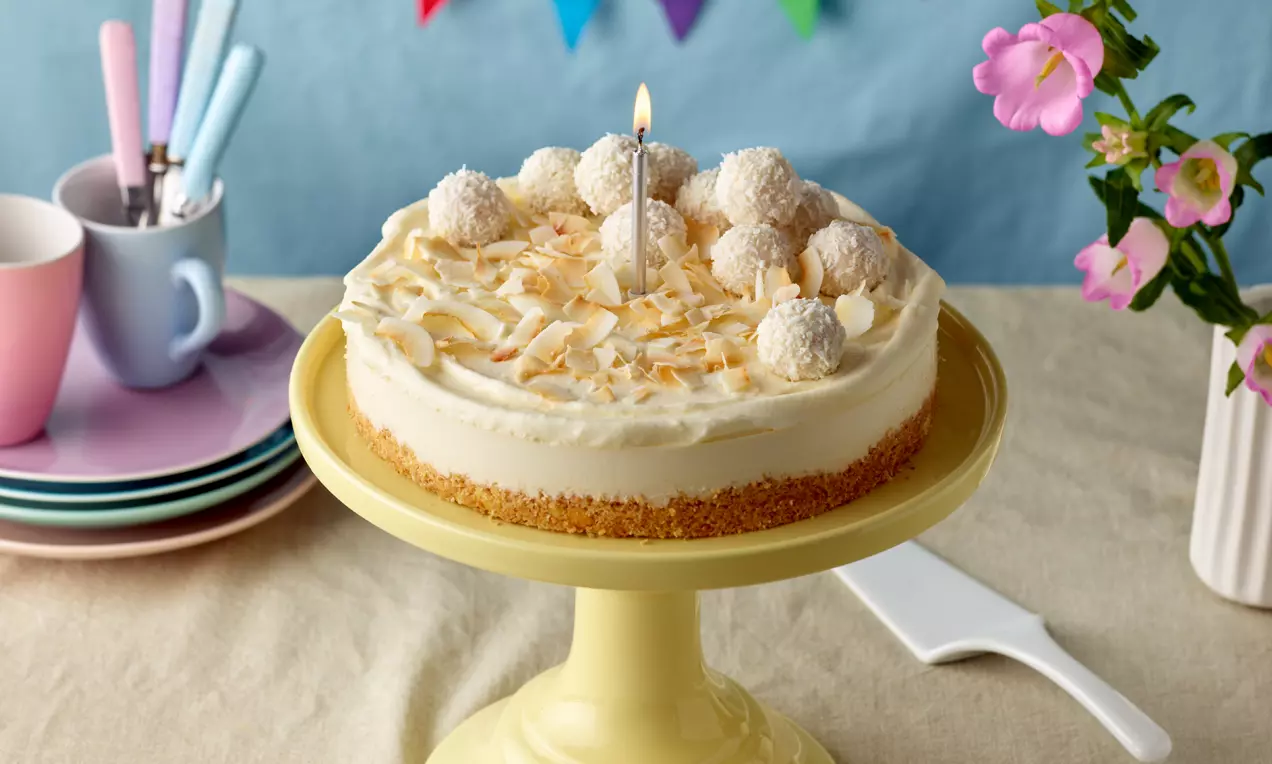 Kokos-Cheesecake mit weißer Frischkäsecreme, goldbraunem Keksboden, Kokosraspeln und Pralinen, dekoriert mit einer Kerze auf gelber Tortenplatte