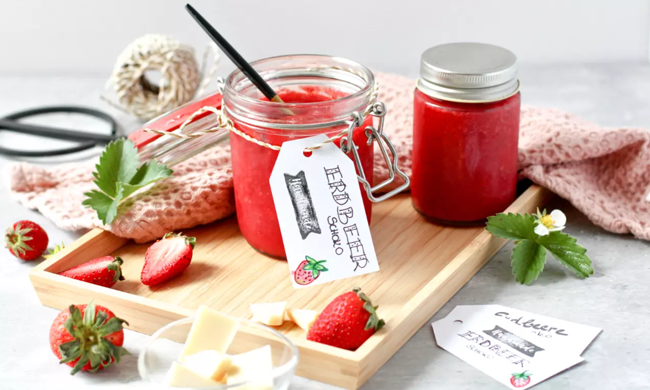 Erdbeermarmelade in einem Glas mit Drahtbügel, daneben weiße Schokowürfel und Erdbeeren auf einem Holzbrett mit rosa Stoff drapiert