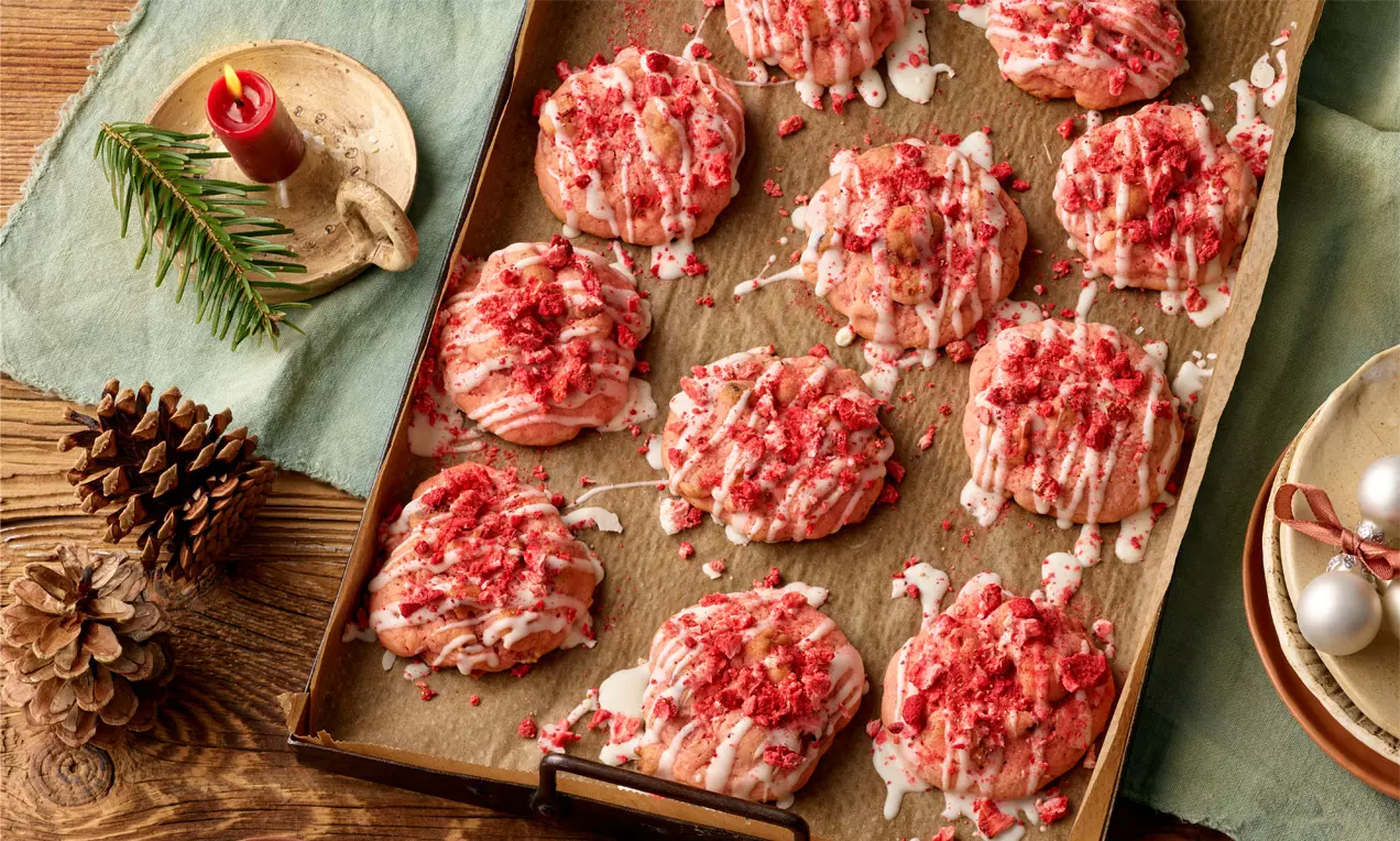Rosafarbene Erdbeer-Cookies mit knusprigen Streuseln und weißer Glasur auf Backpapier auf einem Blech, dekoriert mit Tannenzapfen und Kerze