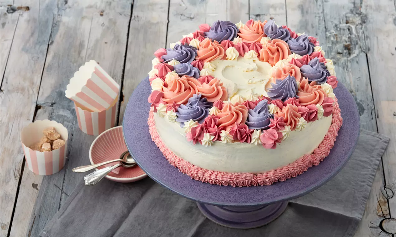 Runde Torte mit weißer Creme, verziert mit fließenden rosa, lila und cremefarbenen Rosetten, präsentiert auf violettem Teller