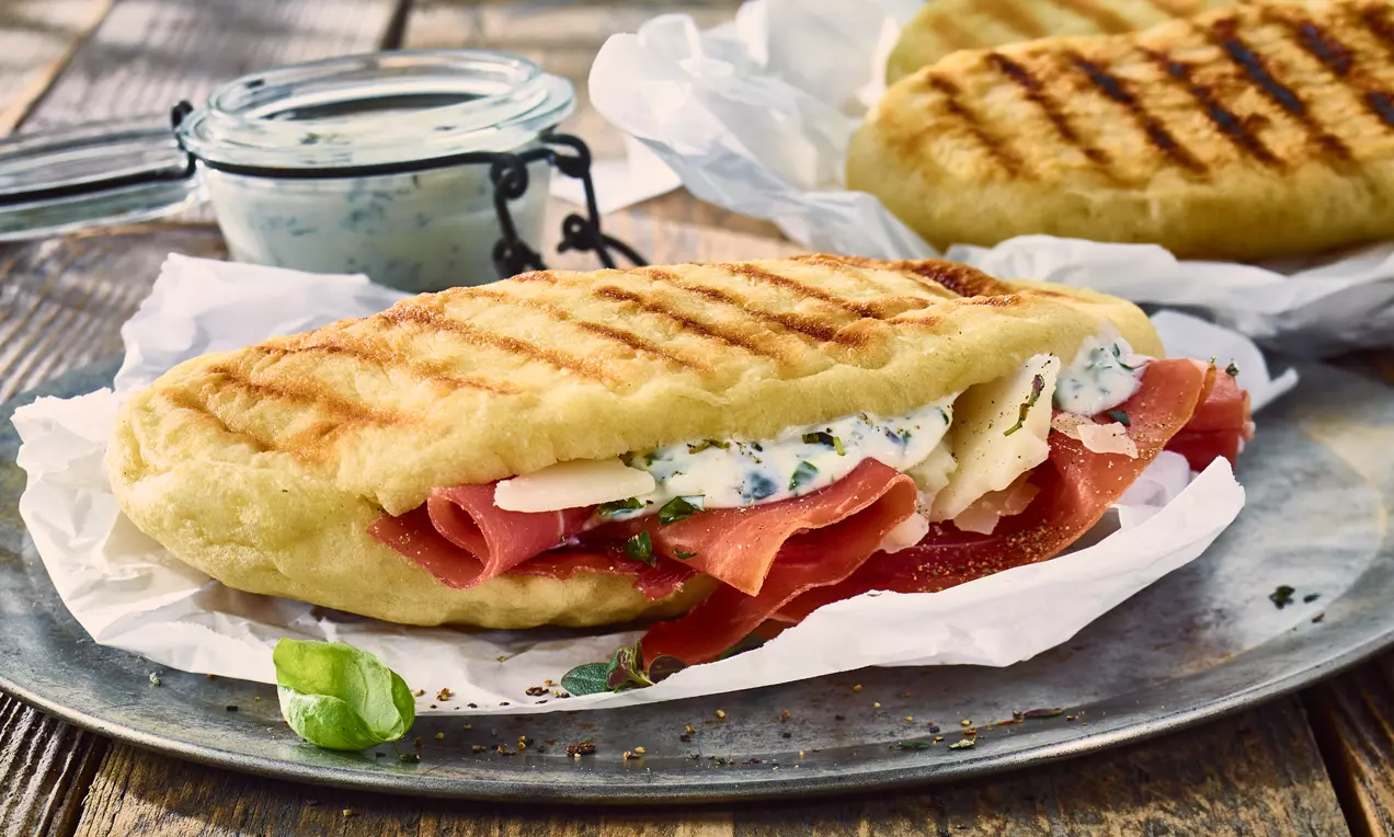 Goldbraune Grillbrote mit zarten Röstaromen, gefüllt mit Schinken, Kräuterdip und Käse auf weißem Papier, daneben ein Glas mit Kräuterdip