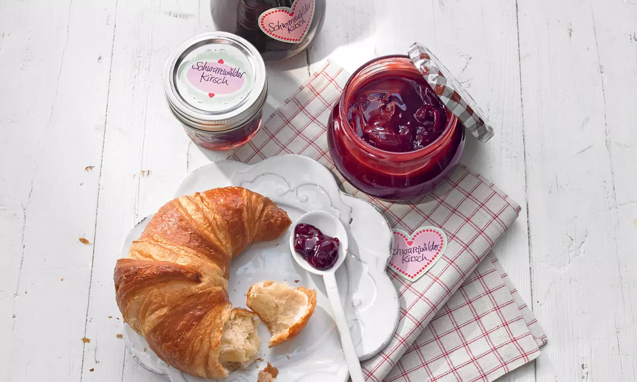 Goldbraunes Croissant und ein weißer Löffel mit dunkelrotem Kirschaufstrich auf einem weißen Teller, daneben offene und geschlossene Gläser