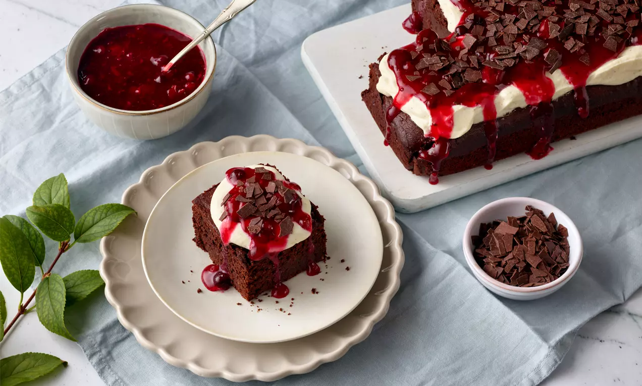 Schokoladiger Brownie mit Sahnehaube und Kirschsoße, bestreut mit dunkler Schokoraspel, auf einem Teller mit frischen Kirschen