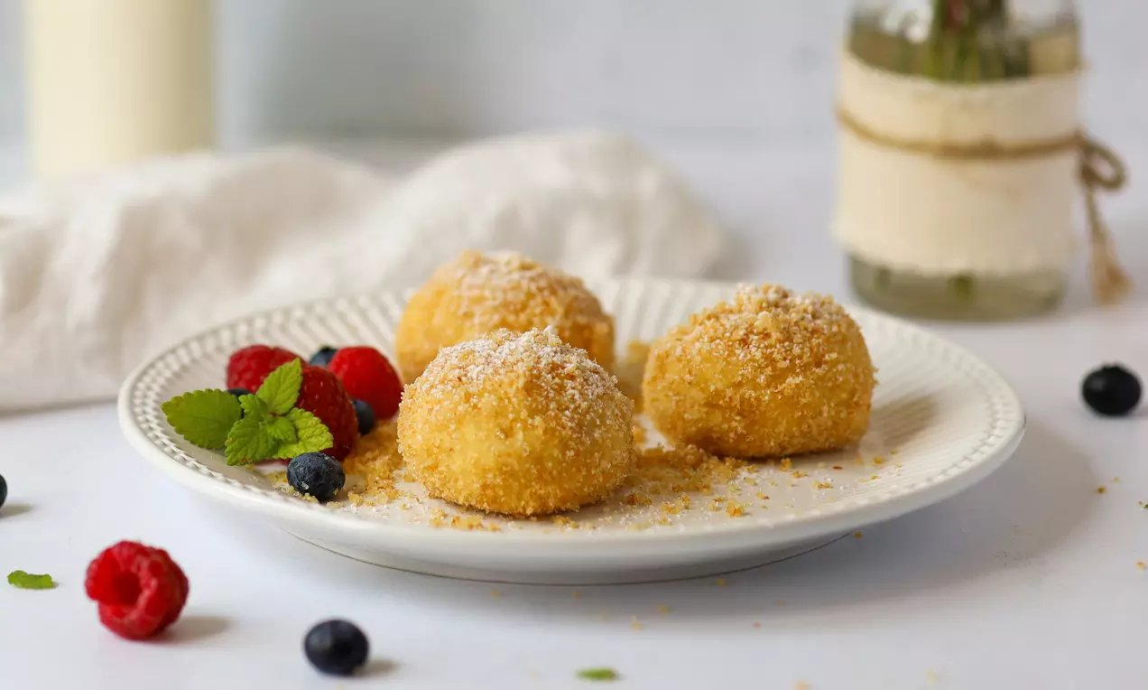 Goldbraune, rundliche Topfenknödel mit feiner Bröselkruste, bestäubt mit Puderzucker, neben frischen Beeren und Minze auf einem weißen Teller