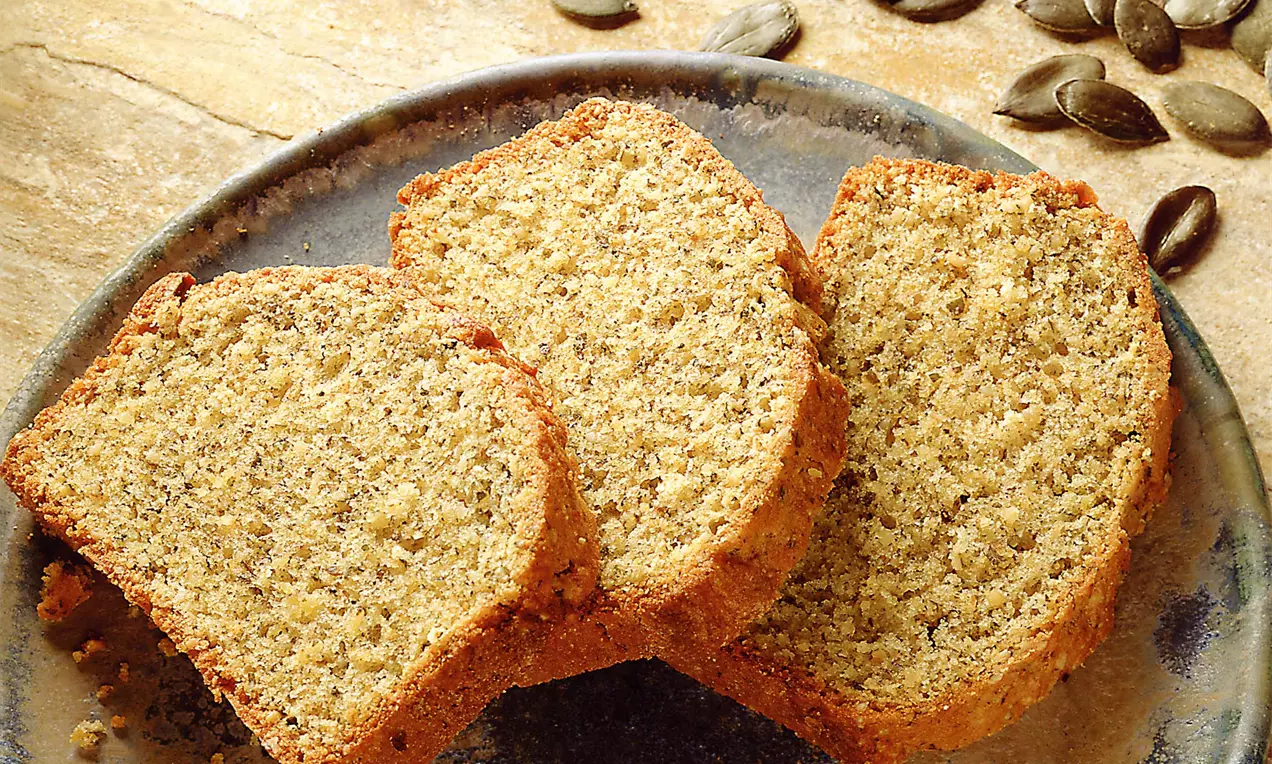 Drei goldbraune Kuchenstücke mit grober, körniger Textur auf dunklem Keramikteller, daneben lose Kürbiskerne auf hellem Untergrund