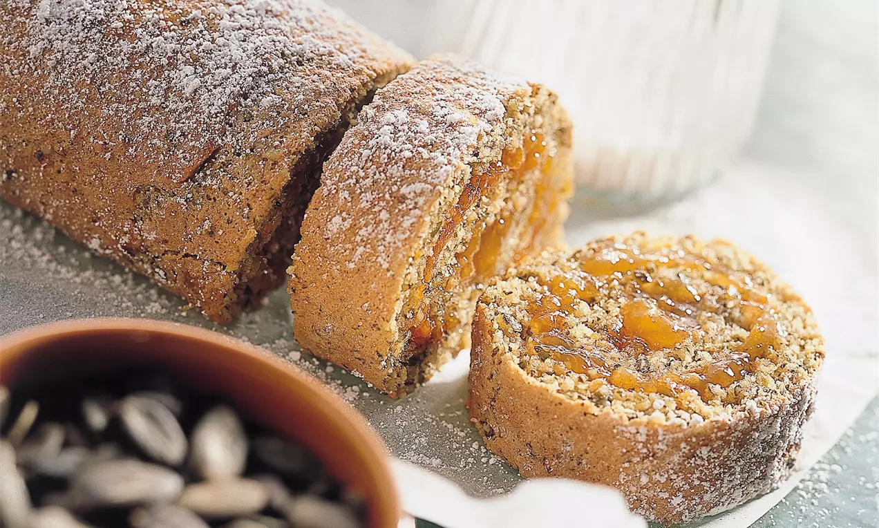 Goldbraune Kürbiskern-Roulade mit sichtbarer Füllung, leicht mit Puderzucker bestäubt, auf Backpapier neben einer Schale Kürbiskerne