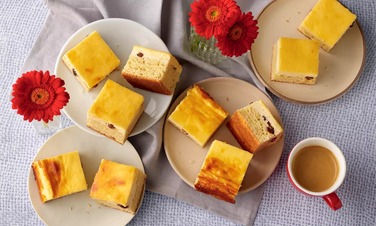 Quadratische Stücke von goldgelbem Topfenfleck mit zarter Textur, auf verschiedenen Tellern arrangiert, umgeben von roten Gerbera und einer Tasse Kaffee