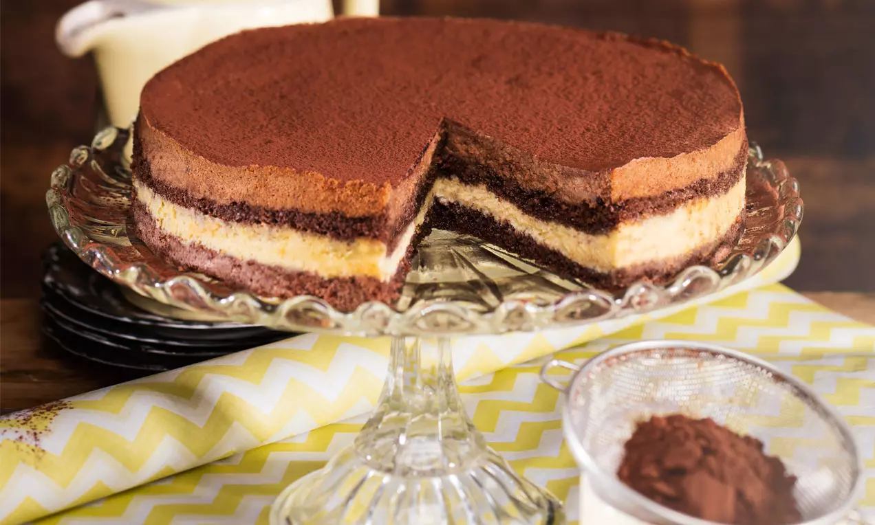 Cheesecake mit Schichten aus weißer und Milchschokolade, bestäubt mit Kakao, auf einer gläsernen Tortenplatte mit gelbem Zickzack-Tuch drapiert