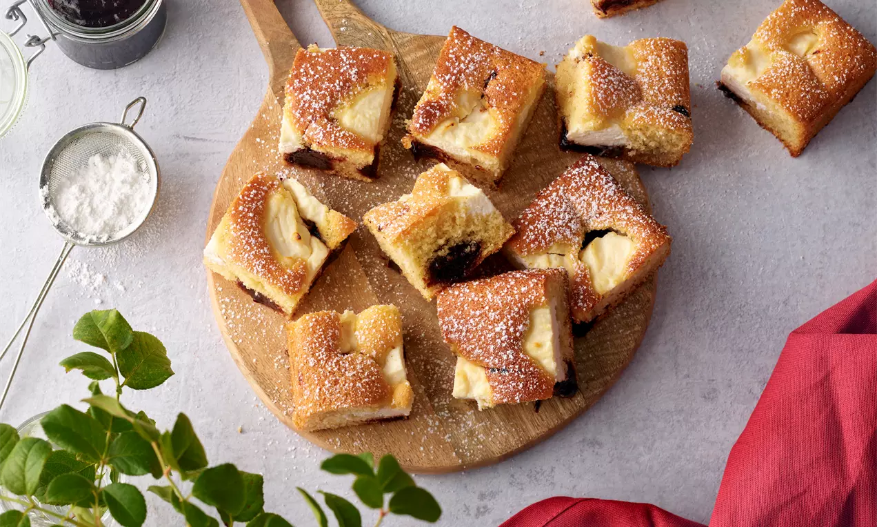 Pflaumen-Ricotta-Kuchen in rechteckigen Stücken mit goldbrauner Kruste und hellem Teig, bestäubt mit Puderzucker, auf einem Holzbrett arrangiert