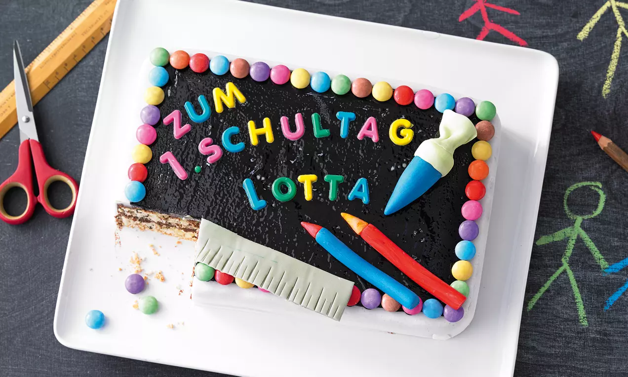Rechteckige Torte mit schwarzer Glasur, bunten Buchstaben, Zuckerstreuseln und Fondant-Dekoration auf weißer Platte mit Kreidetafel-Hintergrund