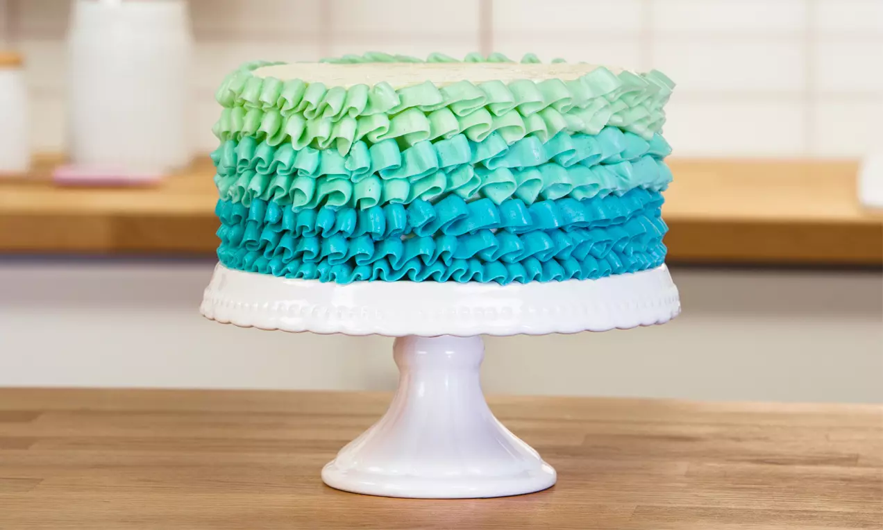 Runde Torte auf weißer Tortenplatte, dekoriert mit mehrfarbigen, wellenförmigen Cremerüschen in Grün- und Blautönen