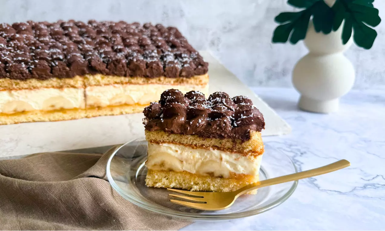 Ein Stück Bananen-Puddingschnitte mit heller Cremefüllung und fluffiger Schokocrème-Schicht auf Glasplatte mit goldener Gabel angerichtet