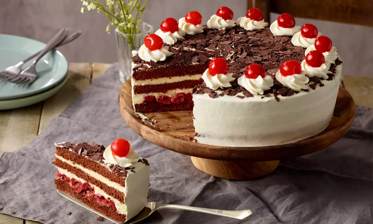 Schwarzwälder Kirschtorte mit weißer Sahne-Ummantelung, Schokoraspeln, Sahnetupfen und roten Kirschen auf einer Holzplatte angerichtet