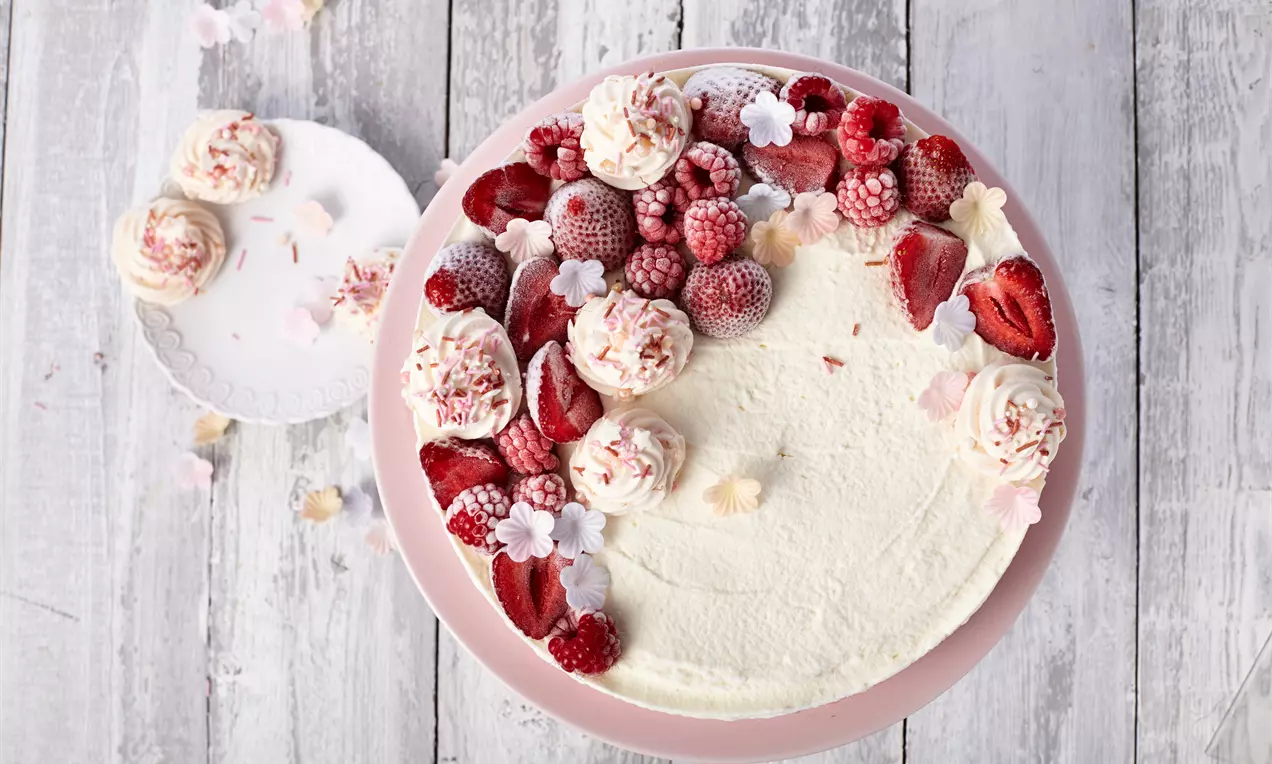 Eistorte mit weißer cremiger Oberfläche, dekoriert mit roten Beeren, Baisertupfen und weißen Blumen auf rosa Tortenplatte