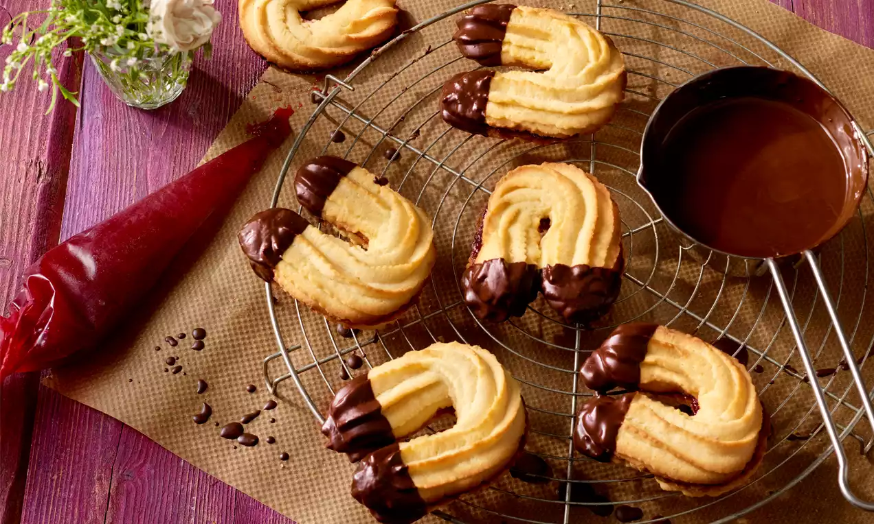 Goldgelbe Linzer Kipferln mit wellenförmiger Struktur, an den Enden in dunkle Schokolade getaucht, auf einem Gitterrost mit Schüssel arrangiert