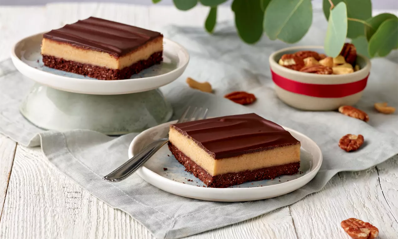 Zwei rechteckige Fitnessschnitten mit dunkler Schokoglasur, heller Creme und braunem Boden auf weißen Tellern mit Dekoration aus Nüssen und Blättern