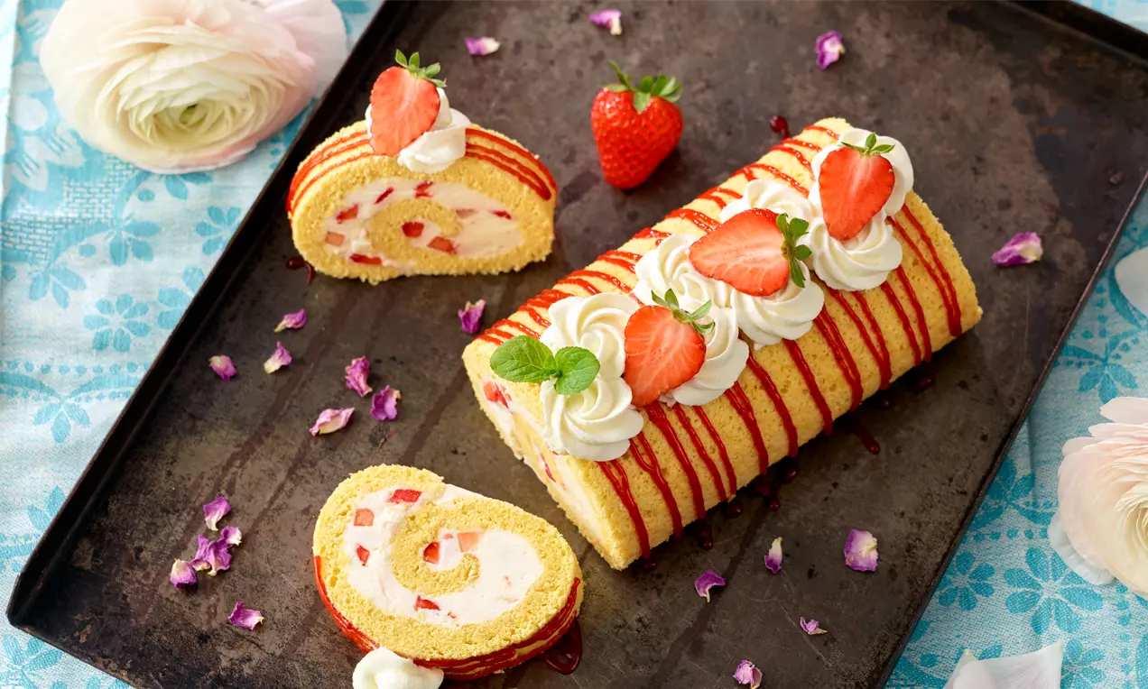 Hellgelbe Biskuitrolle mit roten Erdbeersauce-Streifen, Sahnetupfen, frischen Erdbeeren und Minzblättern auf dunklem Tablett