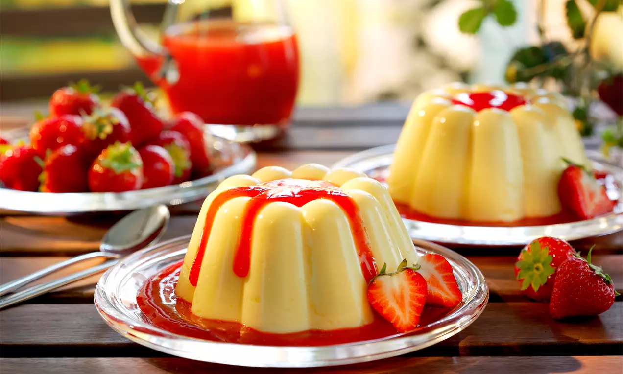 Vanille-Pudding in blassgelber Farbe mit glänzender Erdbeersoße, garniert mit frischen Erdbeeren auf einem gläsernen Dessertteller