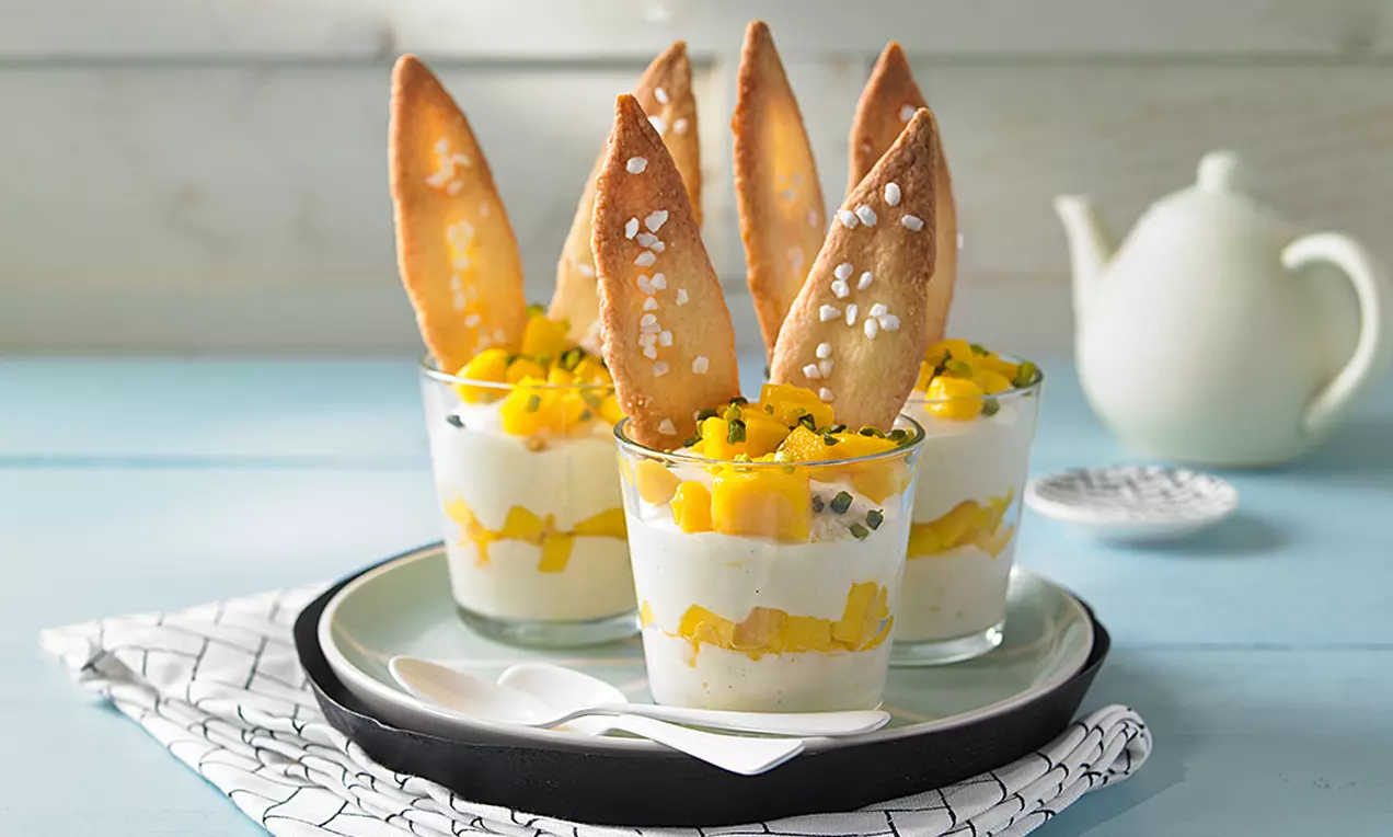 Mango-Mousse in Gläsern, geschichtet mit weißen Cremestreifen, leuchtend gelben Mangostücken und dekorativen, goldbraunen Keksflügeln