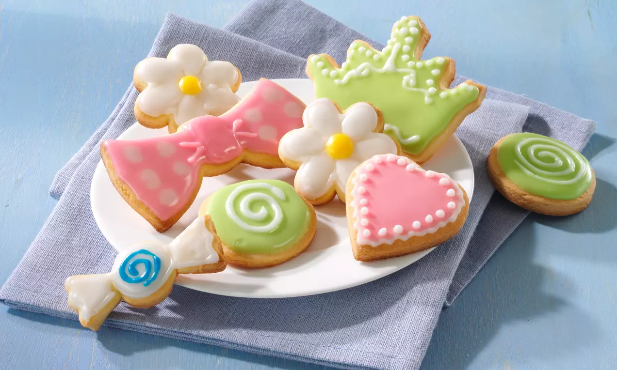 Bunte Butterkekse in Blumen-, Schleifen- und Lutscherform mit glänzendem Zuckerguss auf weißem Teller, drapiert auf einem blauen Stoff