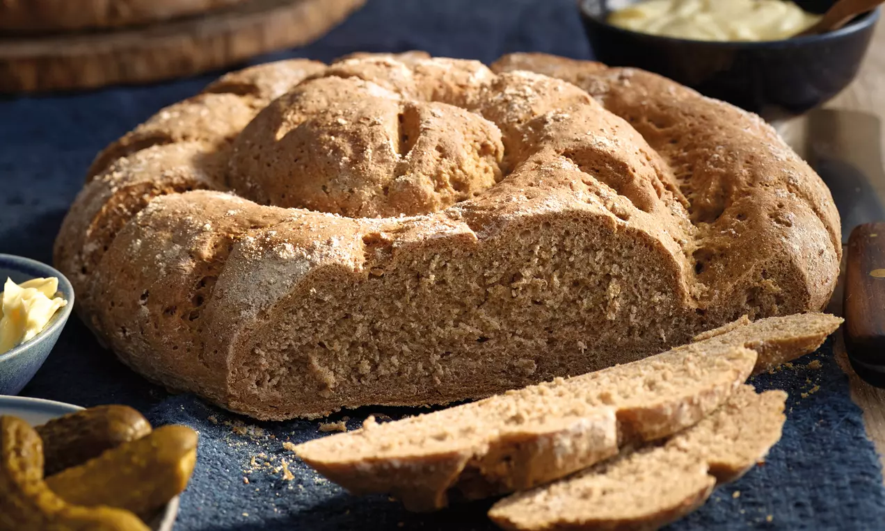 Goldbraunes rundes Landbrot mit grober Kruste und lockerer, körniger Krume, angeschnitten auf dunkler Tischdecke mit Gurkenscheiben und Butter