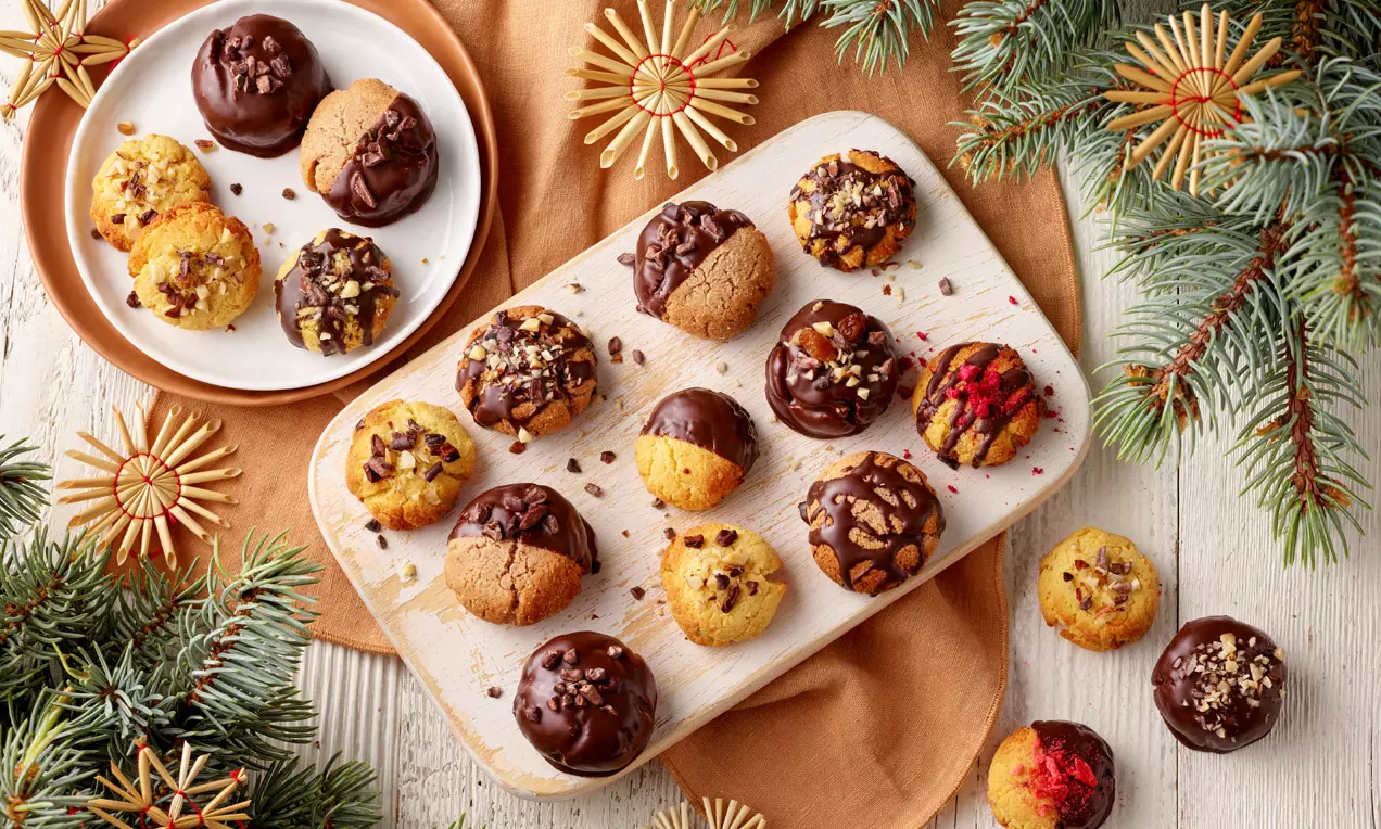 Protein-Cookies in verschiedenen Varianten mit Schokoladenglasur, Nüssen und Beeren auf Holzbrett und Teller, dekoriert mit Tannenzweigen