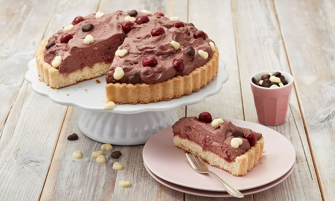 Schokoladen-Schüttelkuchen mit luftiger Creme, getoppt mit Kirschen und Zartbitterschoko-Drops auf weißer Tortenplatte und rosa Teller