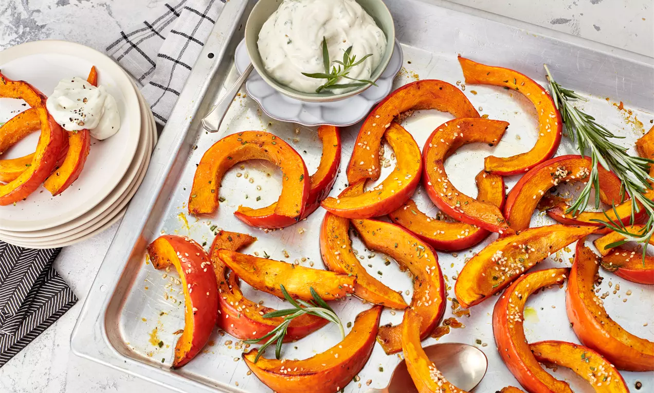 Gebogene orange Kürbisspalten mit goldbrauner Kruste, bestreut mit Gewürzen und Rosmarinzweigen, auf einem silbernen Blech mit Dip-Schälchen