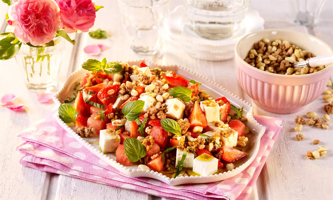 Melonensalat mit roten Erdbeerstücken, weißen Käsewürfeln, frischen Minzblättern und goldbraunem Knusper auf einer weißen Platte mit rosa Stoffservietten