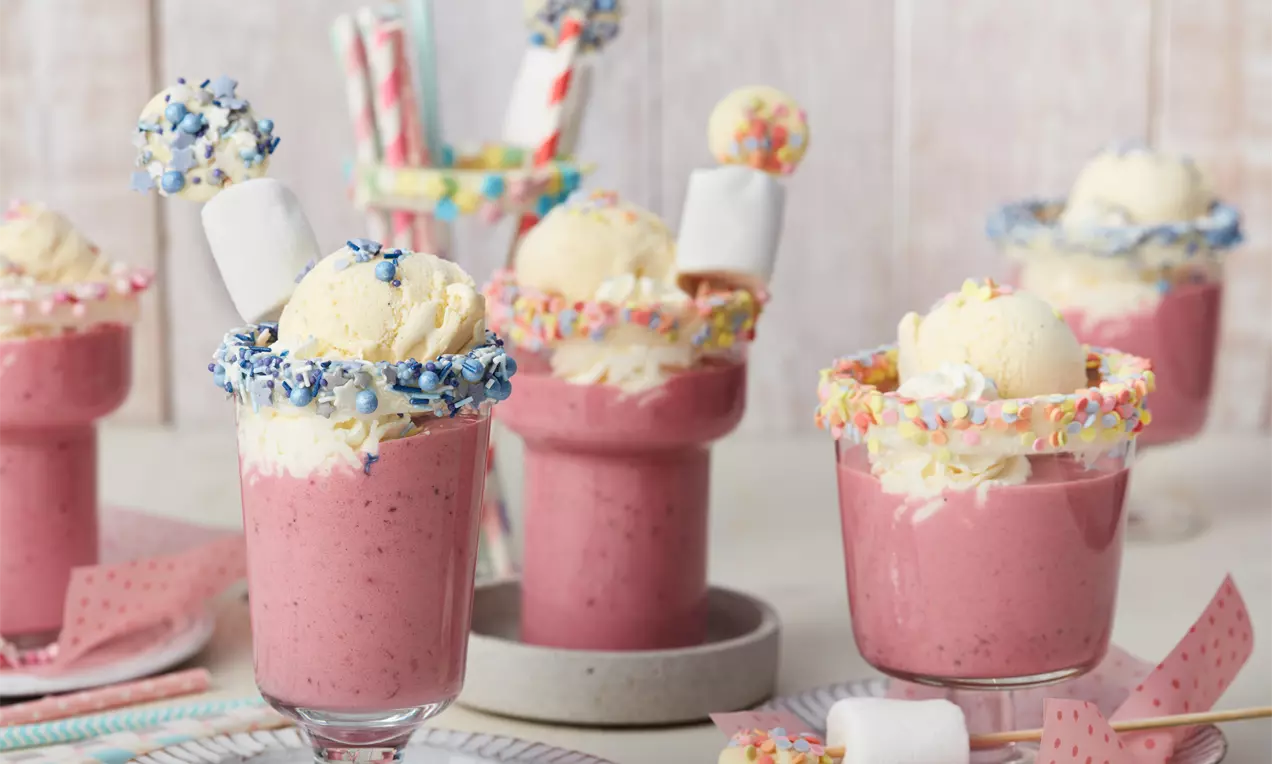 Roter Grütze-Shake in gläsernen Dessertbechern, dekoriert mit bunten Streuseln, Sahne, Eiskugel und Marshmallows auf einem hellen Tischplateau