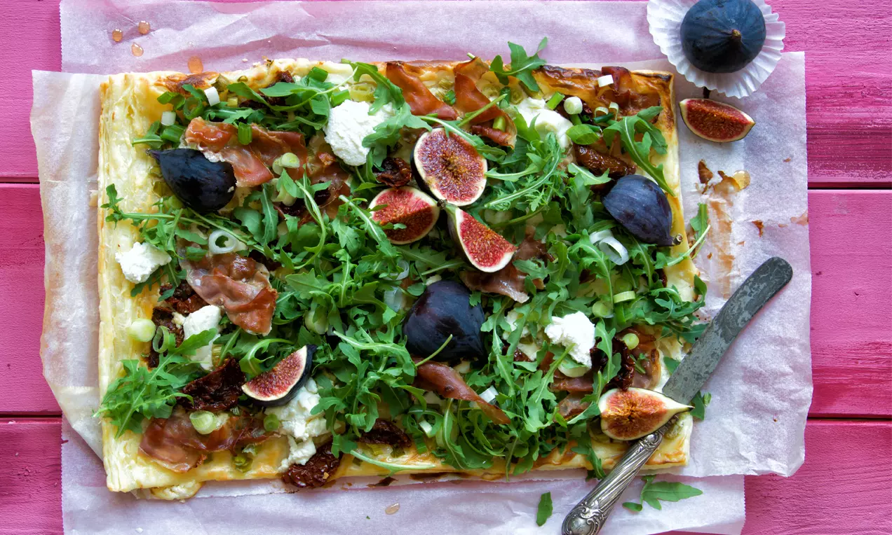 Rechteckiger Blätterteig mit knuspriger goldbrauner Kruste, belegt mit Prosciutto, frischen Feigen, Rucola und weißen Käsehäufchen