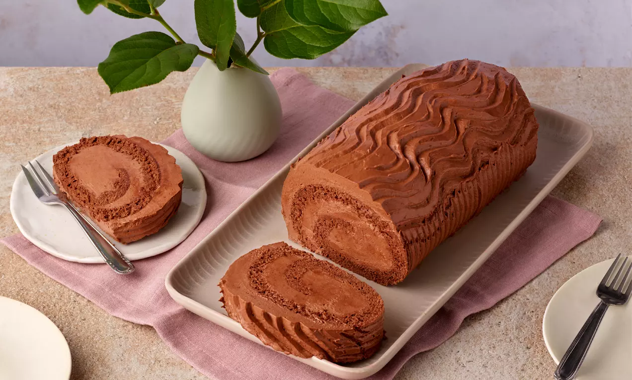 Schoko-Buttercreme-Roulade mit schokoladig-brauner Oberfläche und wellenförmigem Muster, serviert auf einer rechteckigen Platte mit rosa Unterlage