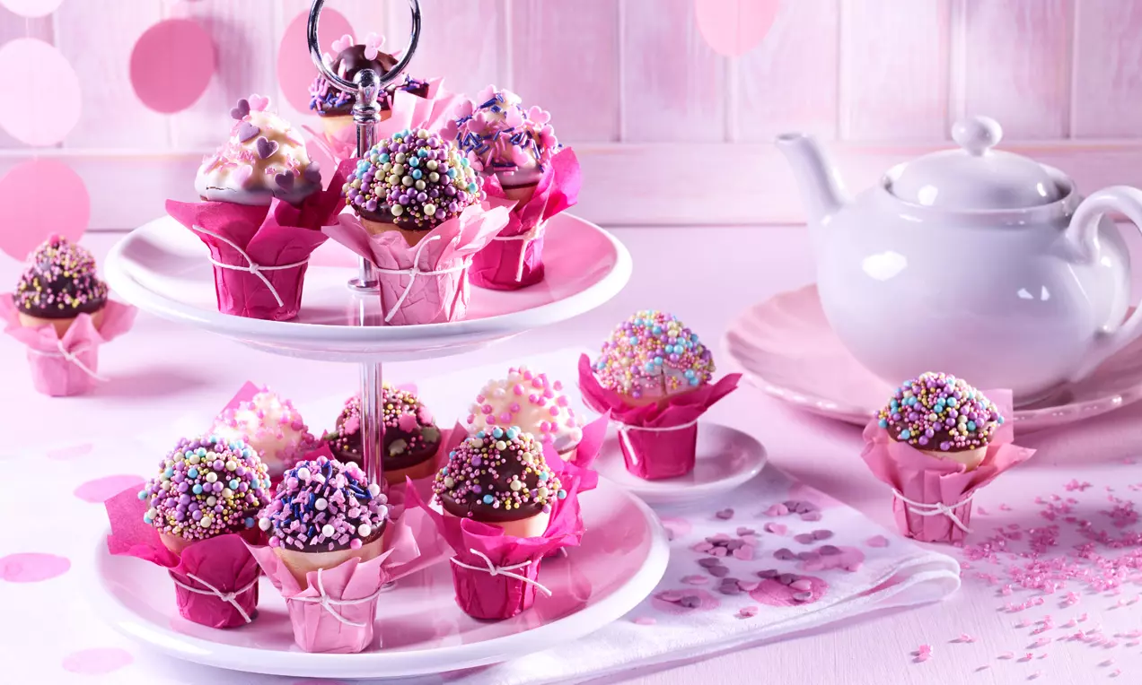 Schokoladenküchlein mit bunter Zuckerstreuselkrone in pinkem Papierförmchen auf weißer Etagere, dekoriert mit Teeservice und rosa Akzenten