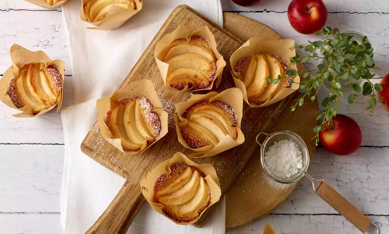 Goldbraune Apfel-Küchlein mit fächerförmig angeordneten Apfelscheiben in braunen Papierförmchen auf rustikalem Holzbrett mit Äpfeln und Dekor