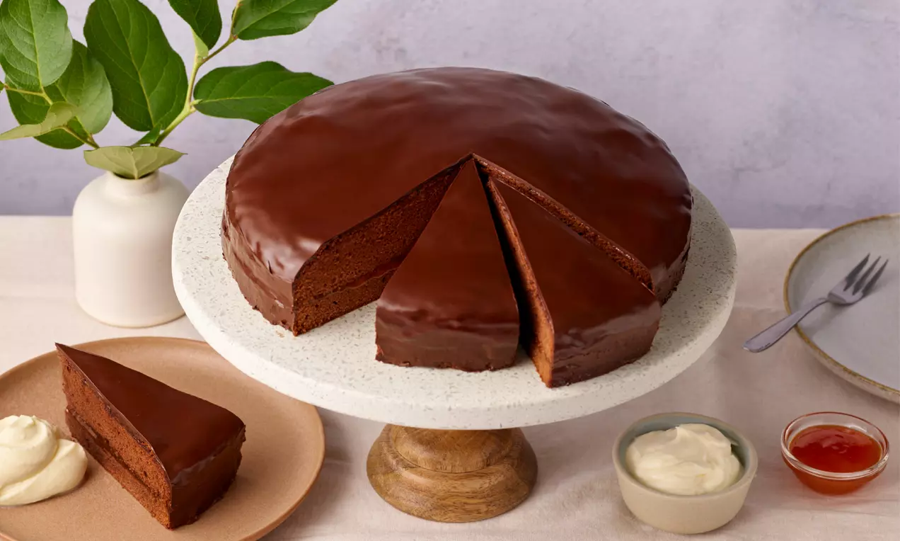 Dunkle, glänzende Sachertorte auf hellem Tortenständer, angeschnitten und serviert mit Schlagobers und Marillenmarmelade auf einem Teller