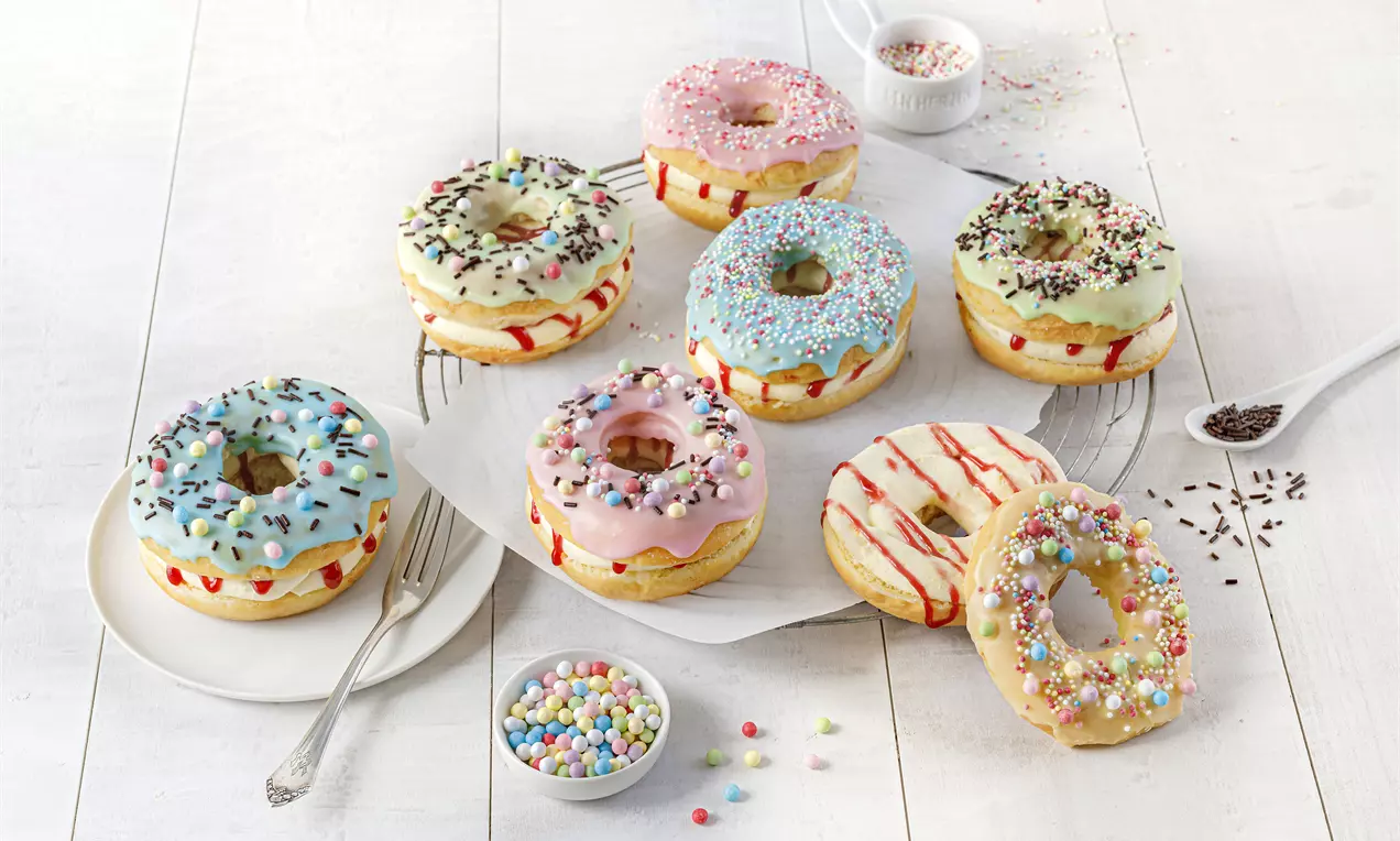 Bunte Donuts mit glänzender Glasur, Puddingfüllung und dekorativen Streuseln auf weißem Teller, flankiert von kleinen Schälchen mit Streudekor