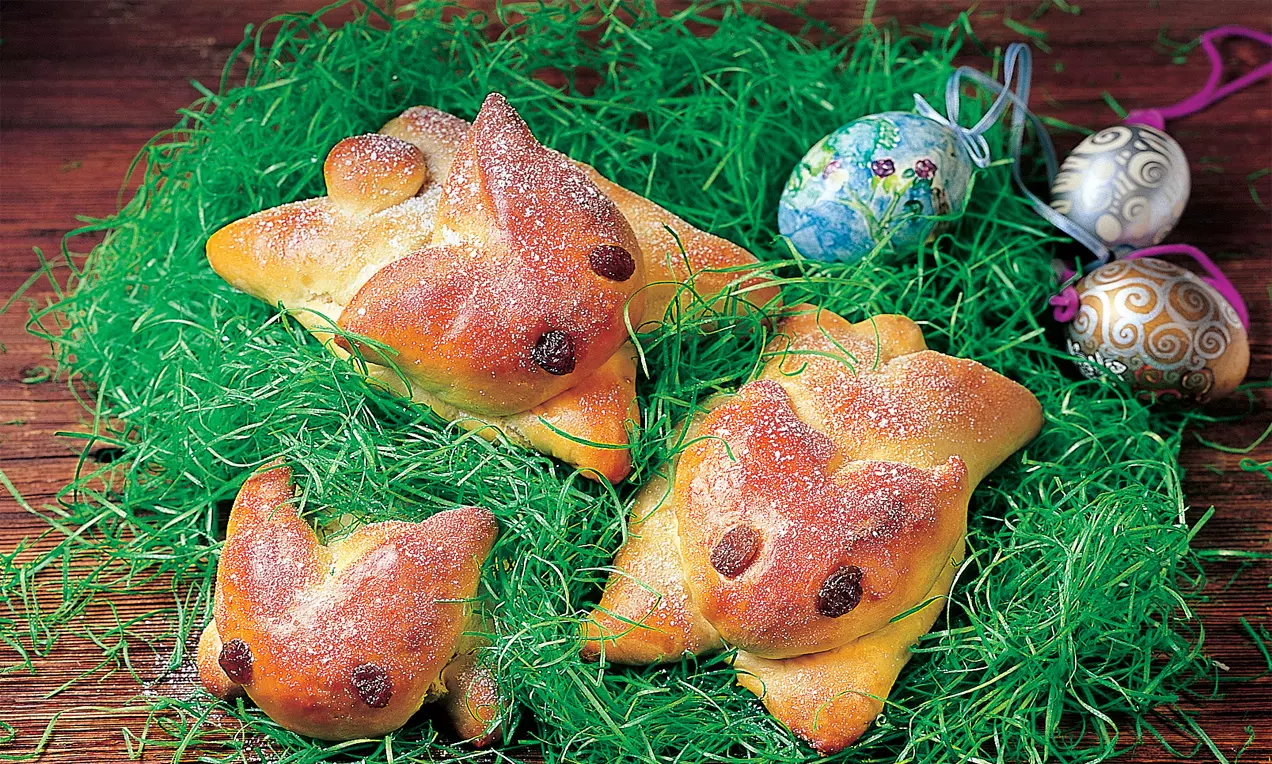 Goldbraune Osterhäschen aus Hefeteig mit Rosinenaugen, leicht bemehlt, auf grünem Ostergras dekoriert, umgeben von bunt verzierten Eiern