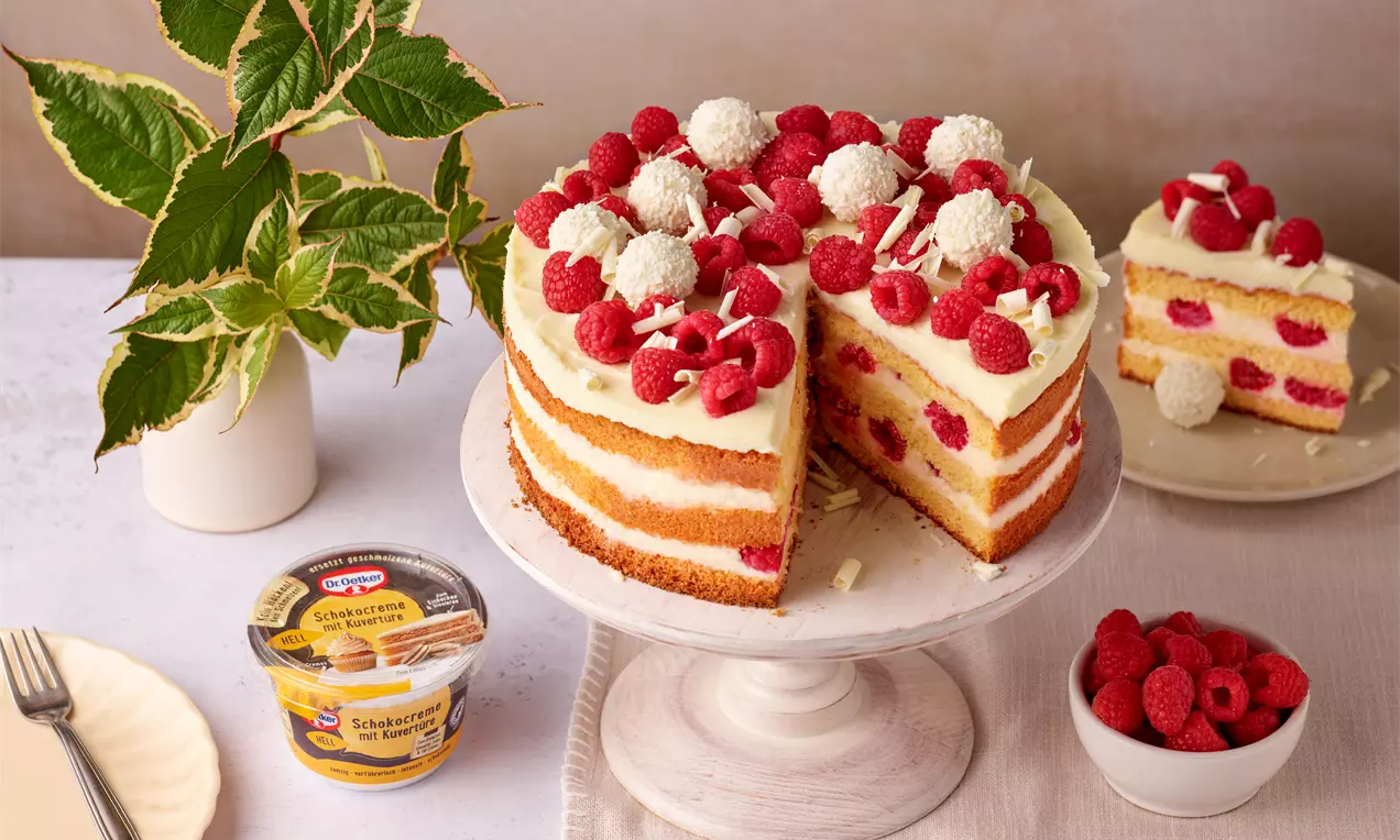 Weiße Schokoladentorte mit Himbeerschichten, dekoriert mit frischen Himbeeren, Raffaello und Schokoraspeln auf einer weißen Kuchenplatte