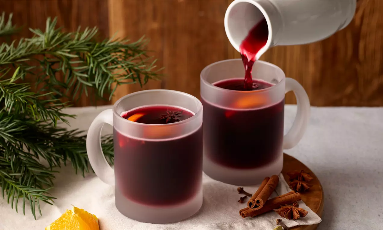 Dunkelroter Glühwein in matten Glastassen, mit Orangenscheiben und Gewürzen auf Holzbrett, daneben Tannenzweige und Zimtstangen