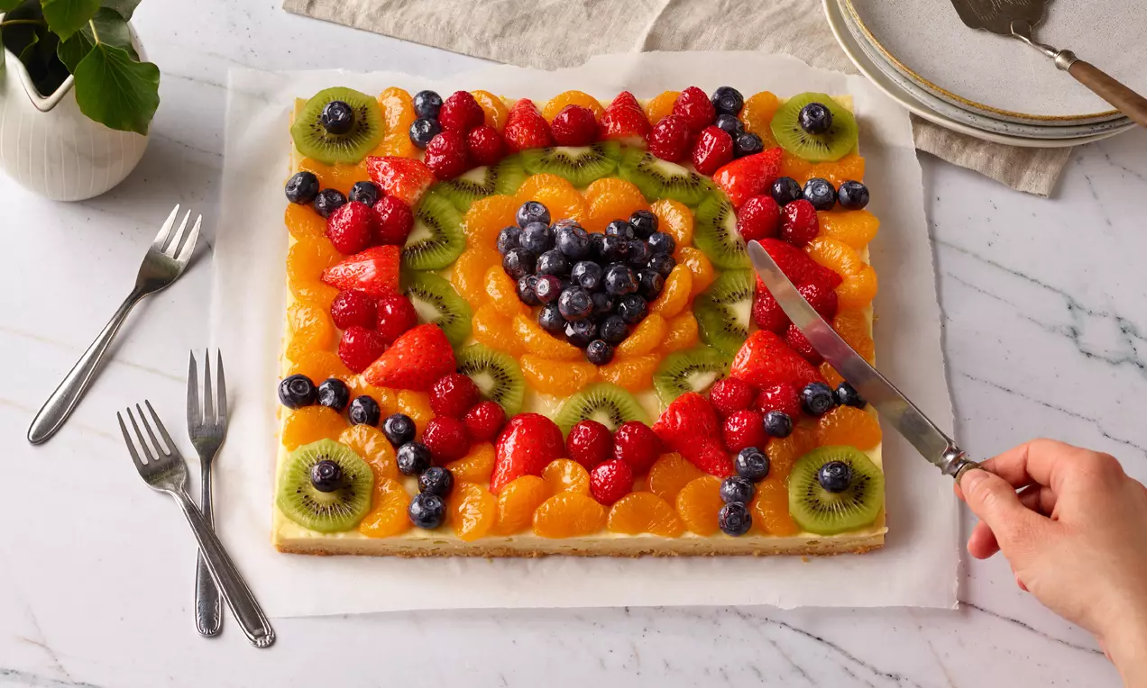 Rechteckiger Pudding-Topfenkuchen mit glänzendem Belag aus Kiwischeiben, Mandarinen, Erdbeeren und Blaubeeren in Herzform auf Backpapier