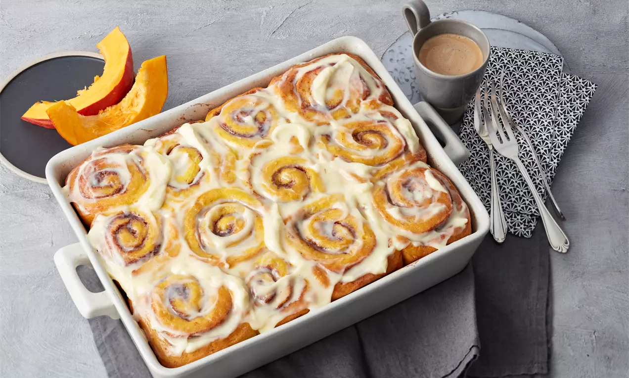 Goldbraune Kürbiszimtschnecken mit spiralförmigem Muster, glasiert mit weißer Creme in einer eckigen weißen Backform auf grauem Untergrund